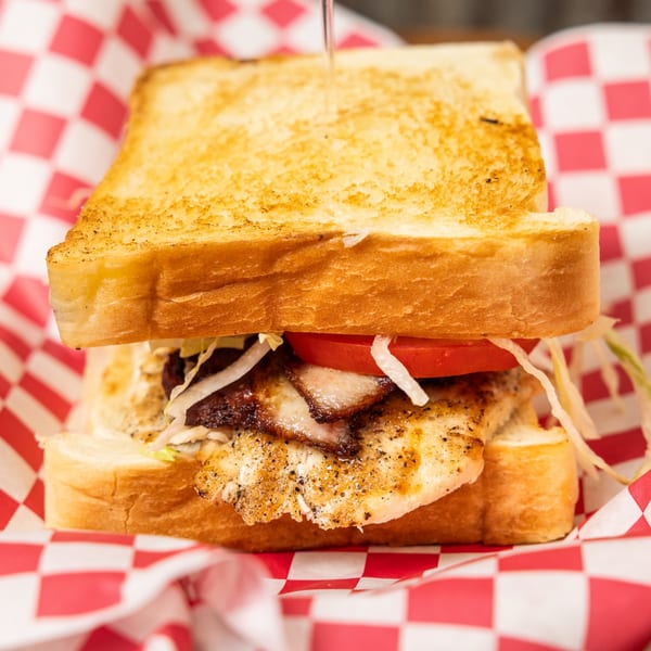 Grilled chicken sandwich with bacon, tomato, and lettuce on toasted bread in a red checkered basket.