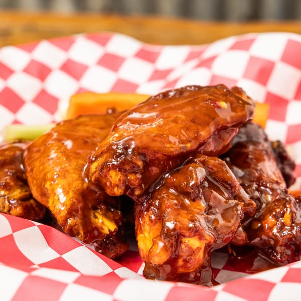 Close-up of Hudson's Grill BBQ chicken wings with celery and carrots in a red checkered basket.