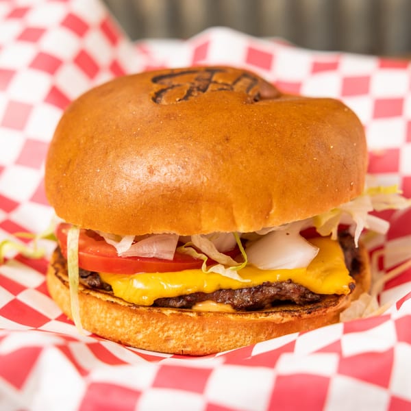 Hudson's Grill burger with cheese, tomato, onion, and lettuce, served in red and white checkered paper.