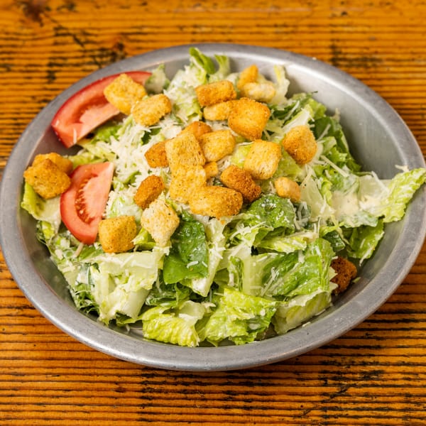 Caesar salad with romaine lettuce, croutons, parmesan cheese, and tomato slices in a metal bowl.