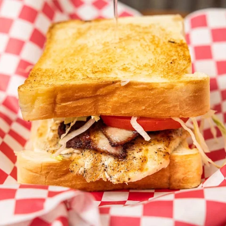 Grilled chicken sandwich with bacon, tomato, and shredded lettuce on toasted bread in a red checkered basket.